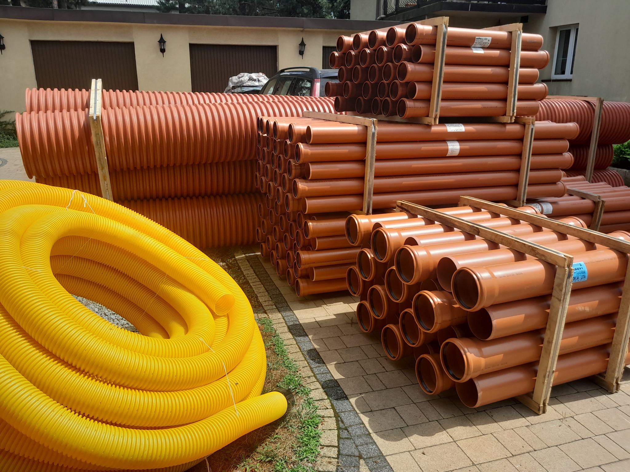 Pomarańczowe rury kanalizacyjne ułożone na paletach obok zwiniętej żółtej rury drenażowej na kostce brukowej.