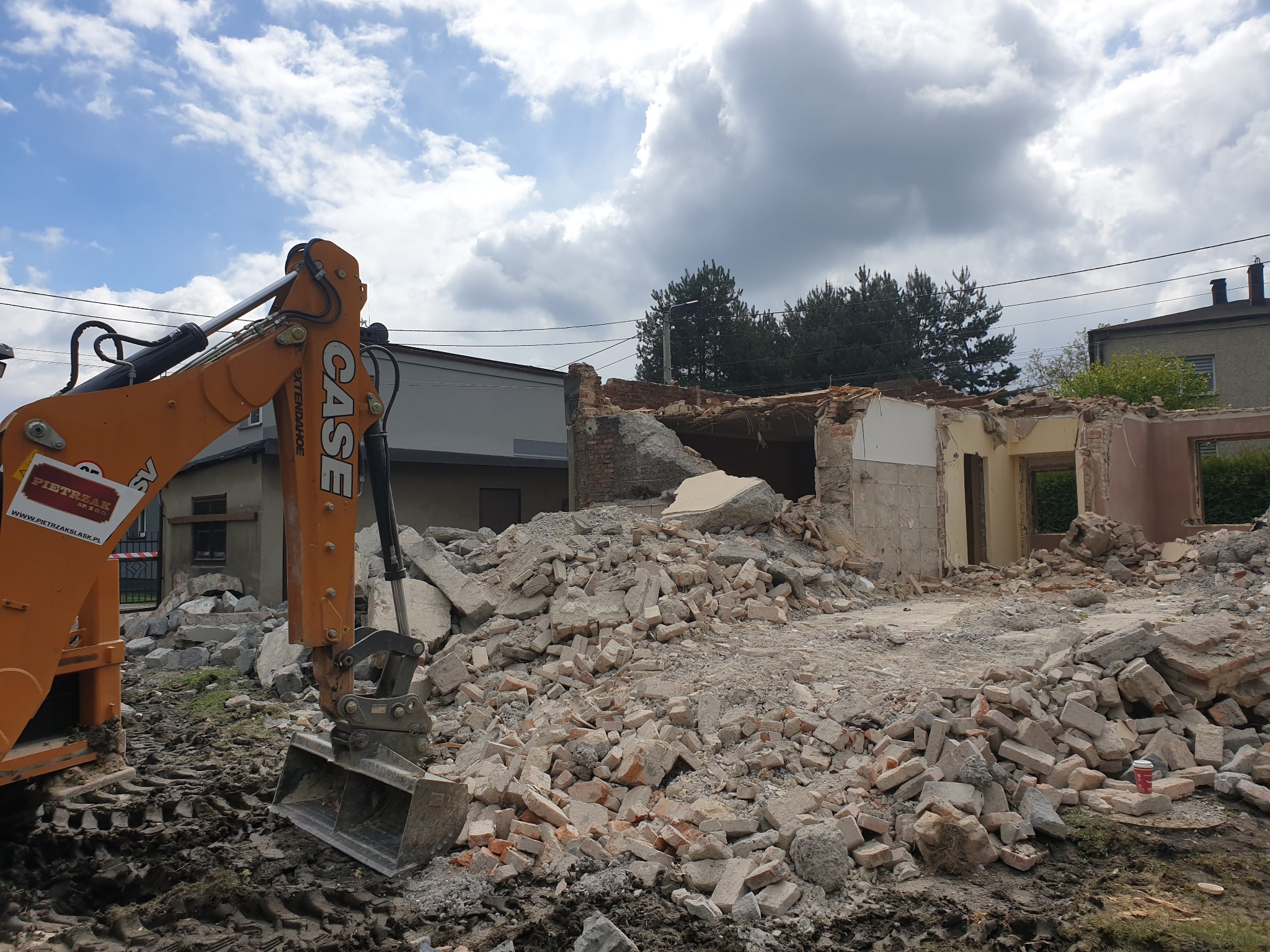Pomarańczowa koparka CASE na tle częściowo zburzonego budynku, sterta gruzu i cegieł, ślady opon w błocie, pochmurne niebo