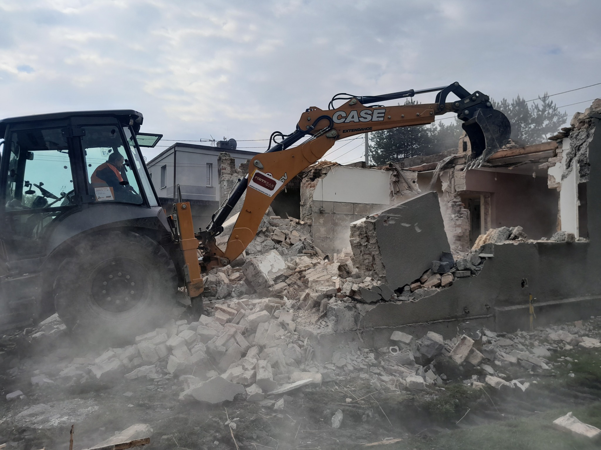 Pomarańczowa koparko-ładowarka Case Extendahoe w trakcie burzenia murowanego budynku, pył i gruz unoszą się w powietrzu, operator w kabinie, w tle budynek mieszkalny.