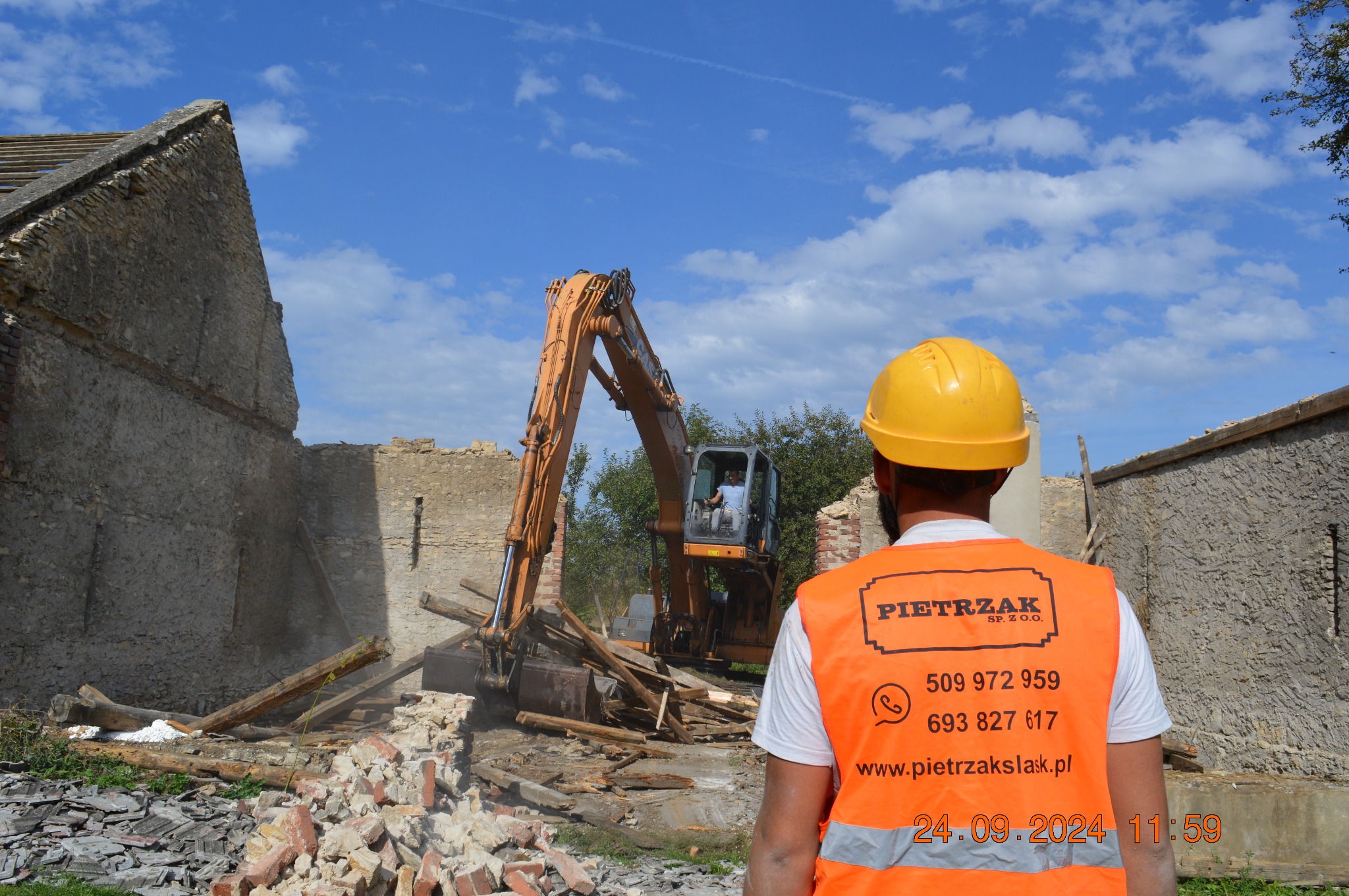 Operator koparki w kabinie, maszyna rozbiera starą kamienną ścianę, w tle błękitne niebo z chmurami, na pierwszym planie pracownik w kamizelce odblaskowej z widocznym logo firmy.