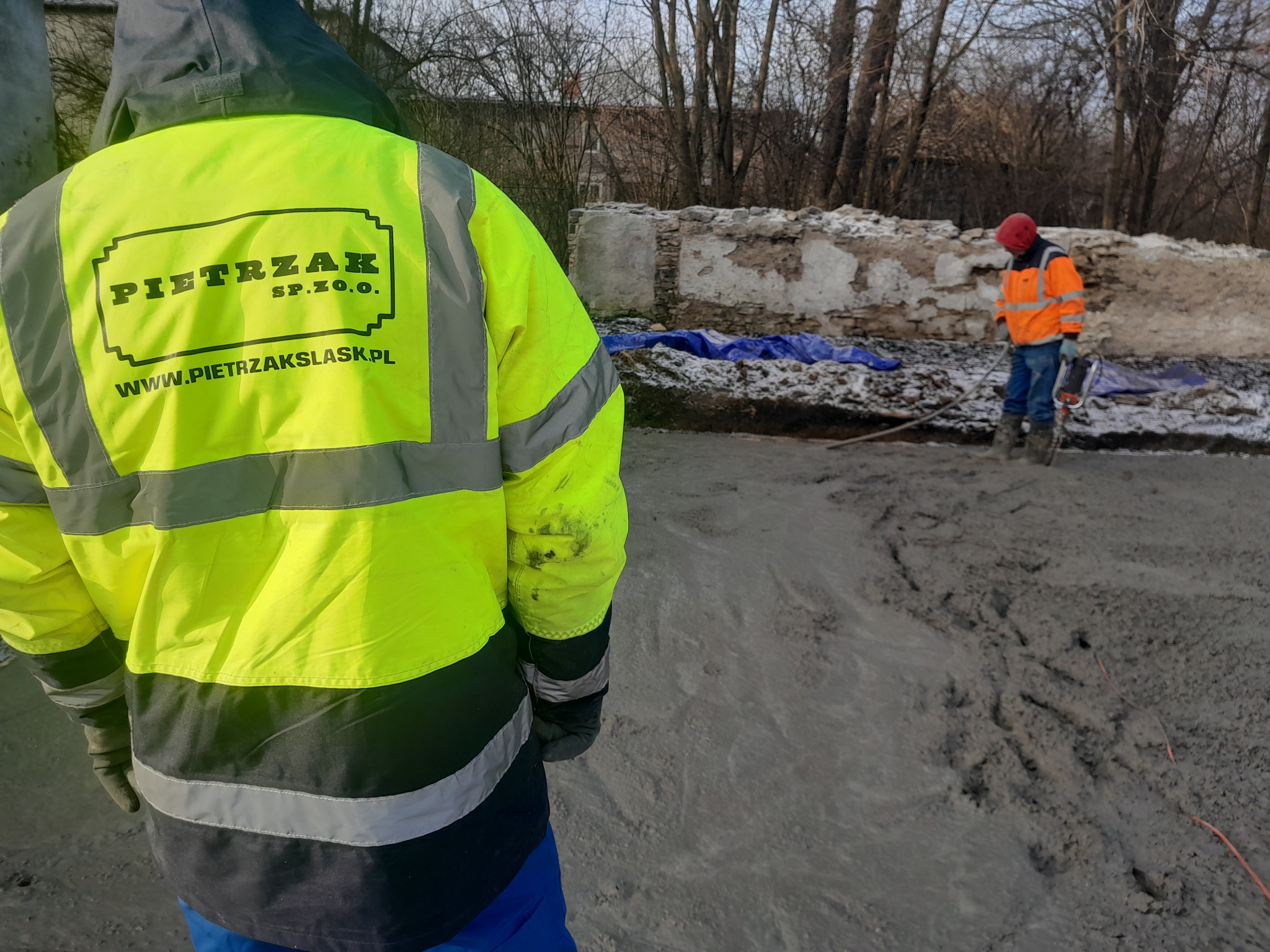 Pracownik w jaskrawej kamizelce z logo firmy PIETRZAK S.P. Z O.O. na tle placu budowy z gruzami i drugim pracownikiem w oddali, wykonującym prace ziemne.