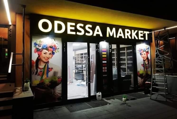 Fasada sklepu Odessa Market z podświetlanym napisem, oklejona grafiką przedstawiającą kobietę w tradycyjnym stroju ukraińskim, widoczne drabiny i akcesoria montażowe.