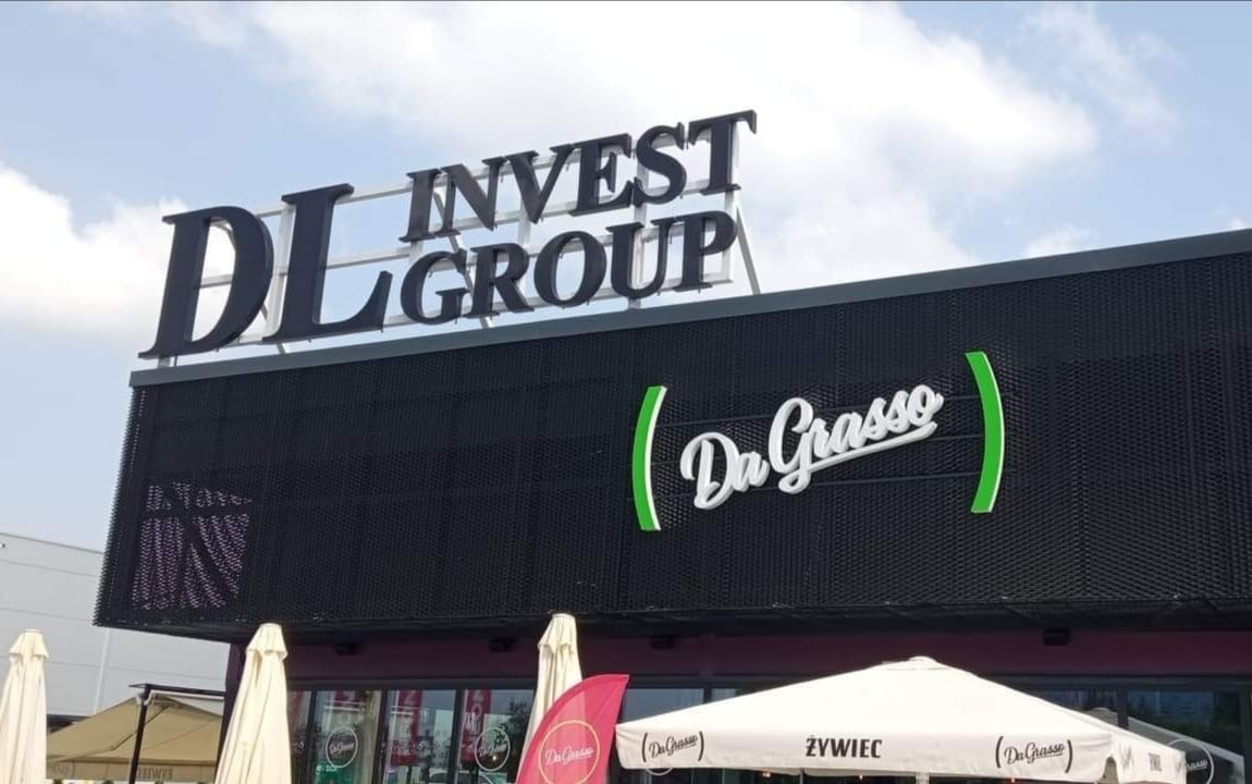 Elewacja budynku z szyldami firmowymi 'DL Invest Group' i 'Da Grasso', widoczne parasole i elementy wystroju restauracji.