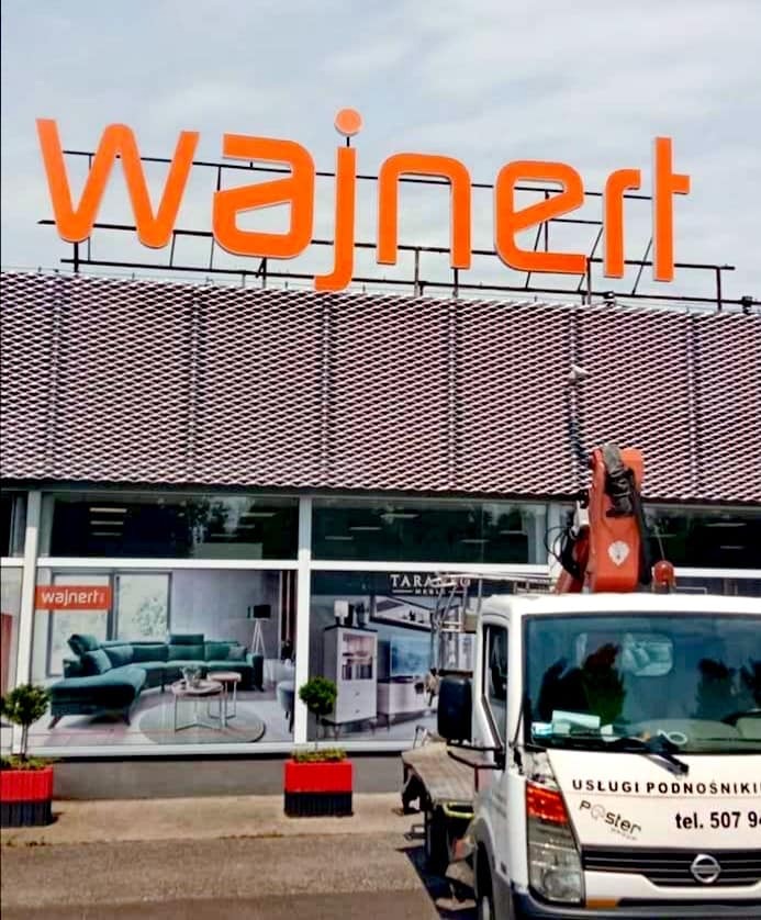 Montaż pomarańczowego napisu 'Wajnert' na dachu budynku z użyciem podnośnika koszowego. W tle widoczne witryny salonu meblowego.