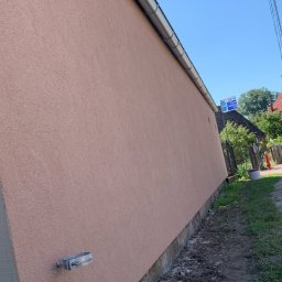 Usługi Budowlane Jakub Jedynak - Ściana budynku po nałożeniu tynku w odcieniach różu, z widoczną metalową rynną i uchwytem na ścianie, w tle zieleń i fragment zabudowań.