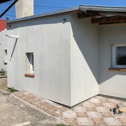 Usługi Budowlane Jakub Jedynak - Świeżo otynkowana elewacja budynku z widocznymi oknami i fragmentem dachu, z ziemią i trawą w dolnej części kadru.