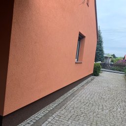 Usługi Budowlane Jakub Jedynak - Fragment pomarańczowej elewacji domu z oknem, widoczny chodnik z kostki brukowej i zieleń w tle.