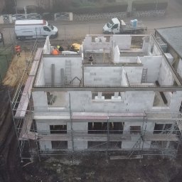Usługi Budowlane Jakub Jedynak - Budowa domu z betonu komórkowego, widok z góry na dwukondygnacyjną konstrukcję w trakcie wznoszenia ścian, z widocznymi rusztowaniami, pracownikami w odblaskowych kamizelkach oraz sprzętem...