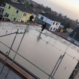 Usługi Budowlane Jakub Jedynak - Wylewanie betonu na strop budynku jednorodzinnego, widoczne szalunki, stalowe pręty zbrojeniowe oraz pracownicy w żółtych gumowcach wygładzający powierzchnię betonu.