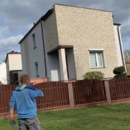 Dwukondygnacyjny dom z elewacją z kamienia dekoracyjnego. Na pierwszym planie trawnik i drewniane ogrodzenie. Widoczna osoba w roboczym ubraniu.