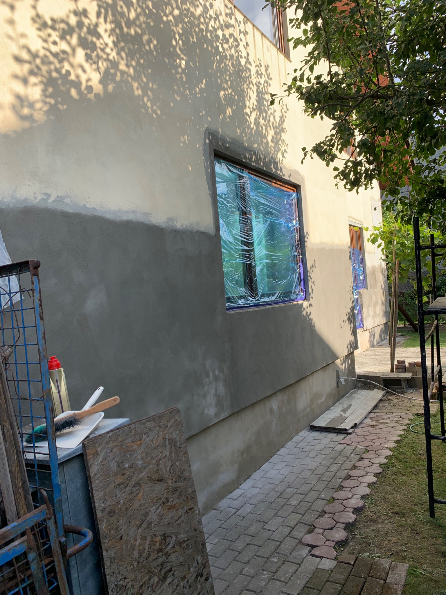 Elewacja budynku w trakcie prac tynkarskich, okna zabezpieczone folią, widoczne narzędzia pracy i cień drzew na ścianie.
