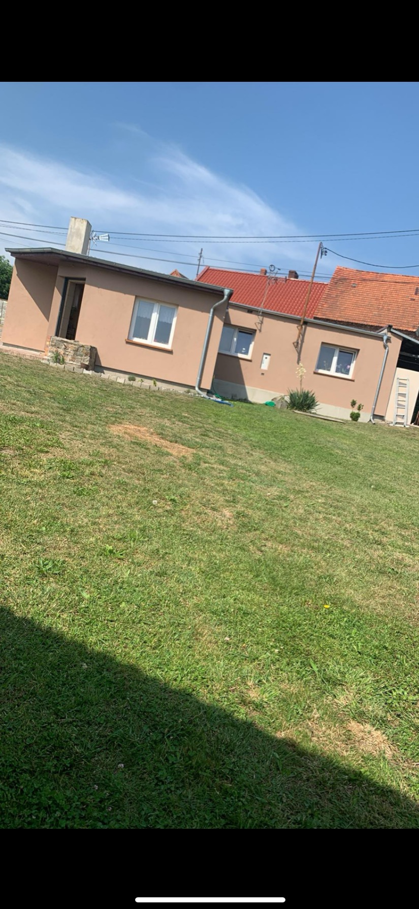 Niski dom z beżową elewacją, czerwonym dachem i białymi oknami, widziany z trawnika w słoneczny dzień.