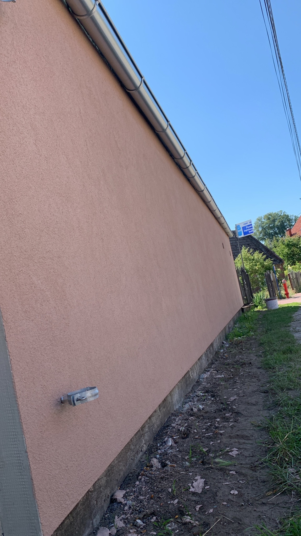 Ściana budynku po nałożeniu tynku w odcieniach różu, z widoczną metalową rynną i uchwytem na ścianie, w tle zieleń i fragment zabudowań.