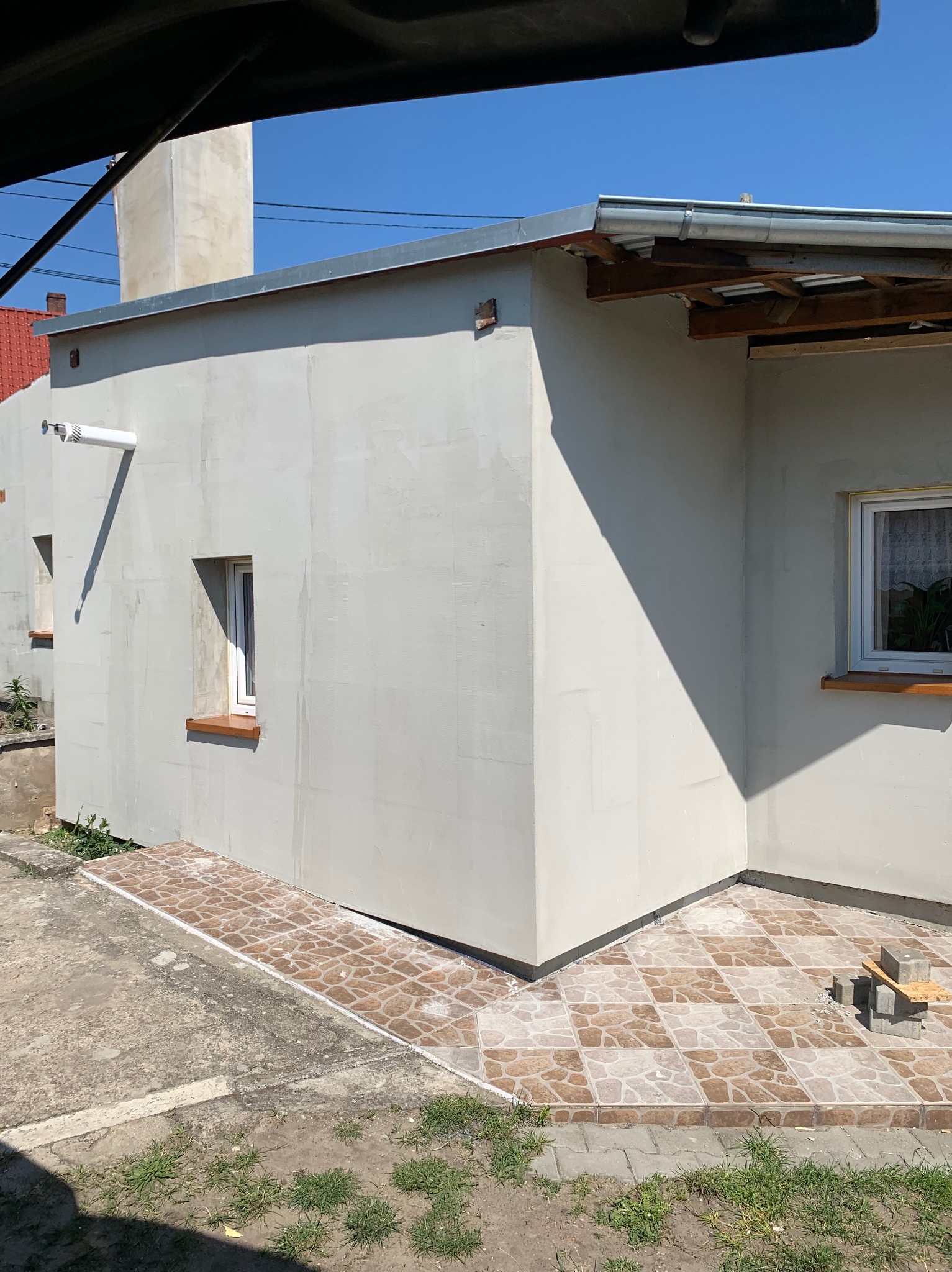 Świeżo otynkowana elewacja budynku z widocznymi oknami i fragmentem dachu, z ziemią i trawą w dolnej części kadru.