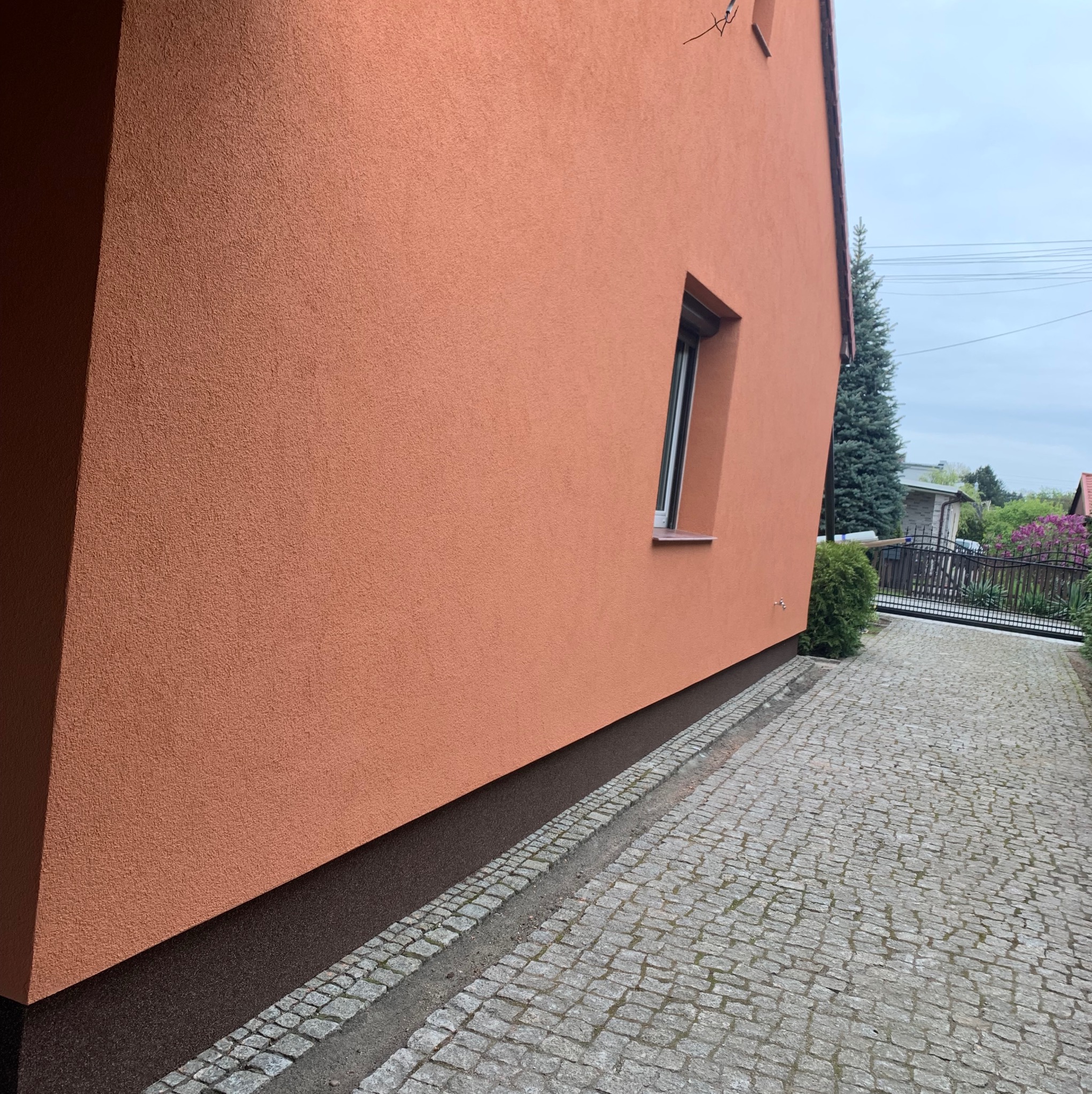 Fragment pomarańczowej elewacji domu z oknem, widoczny chodnik z kostki brukowej i zieleń w tle.