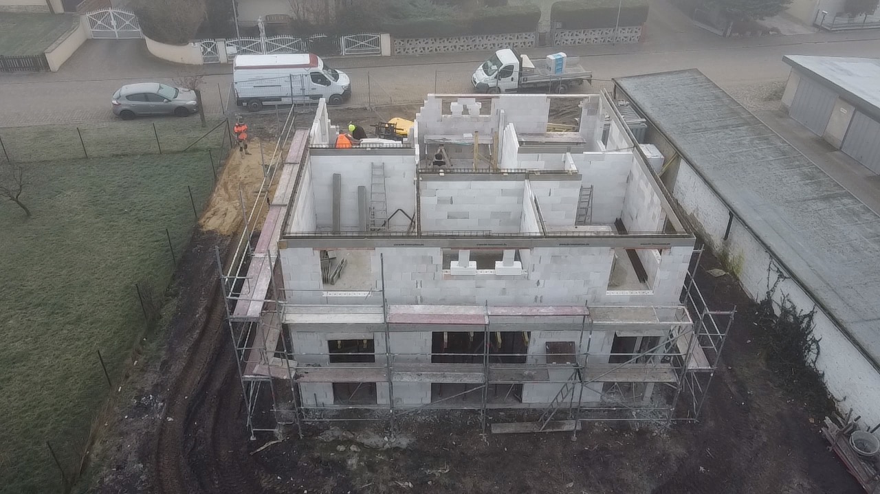 Budowa domu z betonu komórkowego, widok z góry na dwukondygnacyjną konstrukcję w trakcie wznoszenia ścian, z widocznymi rusztowaniami, pracownikami w odblaskowych kamizelkach oraz sprzętem...