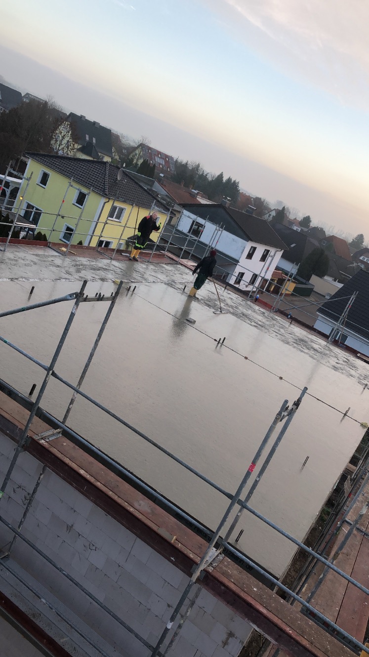 Wylewanie betonu na strop budynku jednorodzinnego, widoczne szalunki, stalowe pręty zbrojeniowe oraz pracownicy w żółtych gumowcach wygładzający powierzchnię betonu.