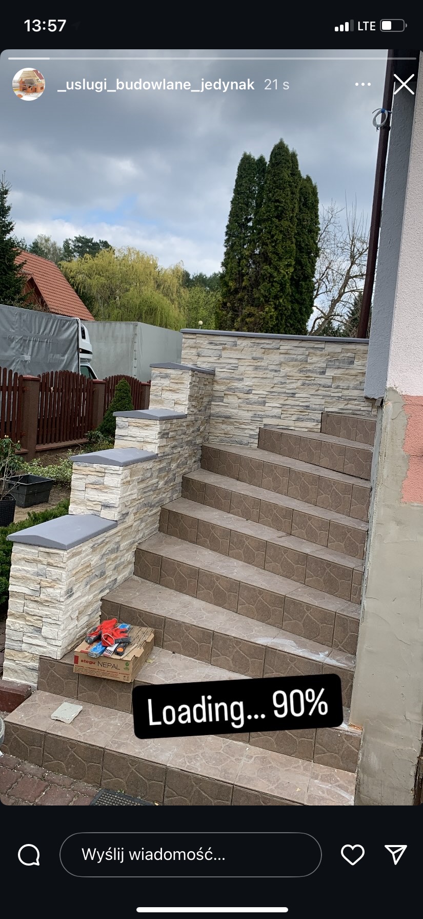 Wykończone schody zewnętrzne z brązowych płytek i kamienną okładziną w odcieniach beżu. Na dole karton z narzędziami. Widok na dom i zieleń w tle.