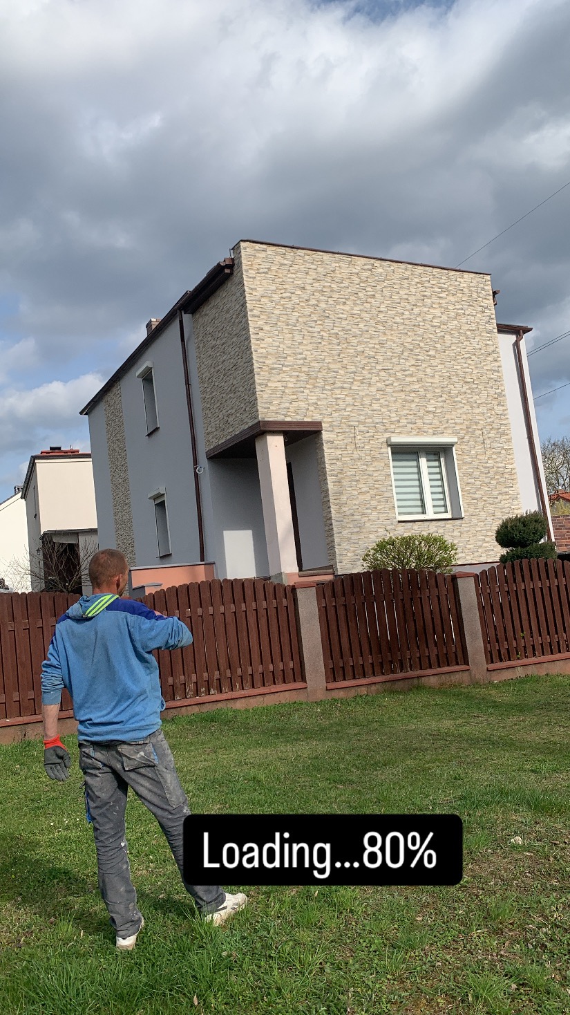 Dwukondygnacyjny dom z elewacją z kamienia dekoracyjnego. Na pierwszym planie trawnik i drewniane ogrodzenie. Widoczna osoba w roboczym ubraniu.