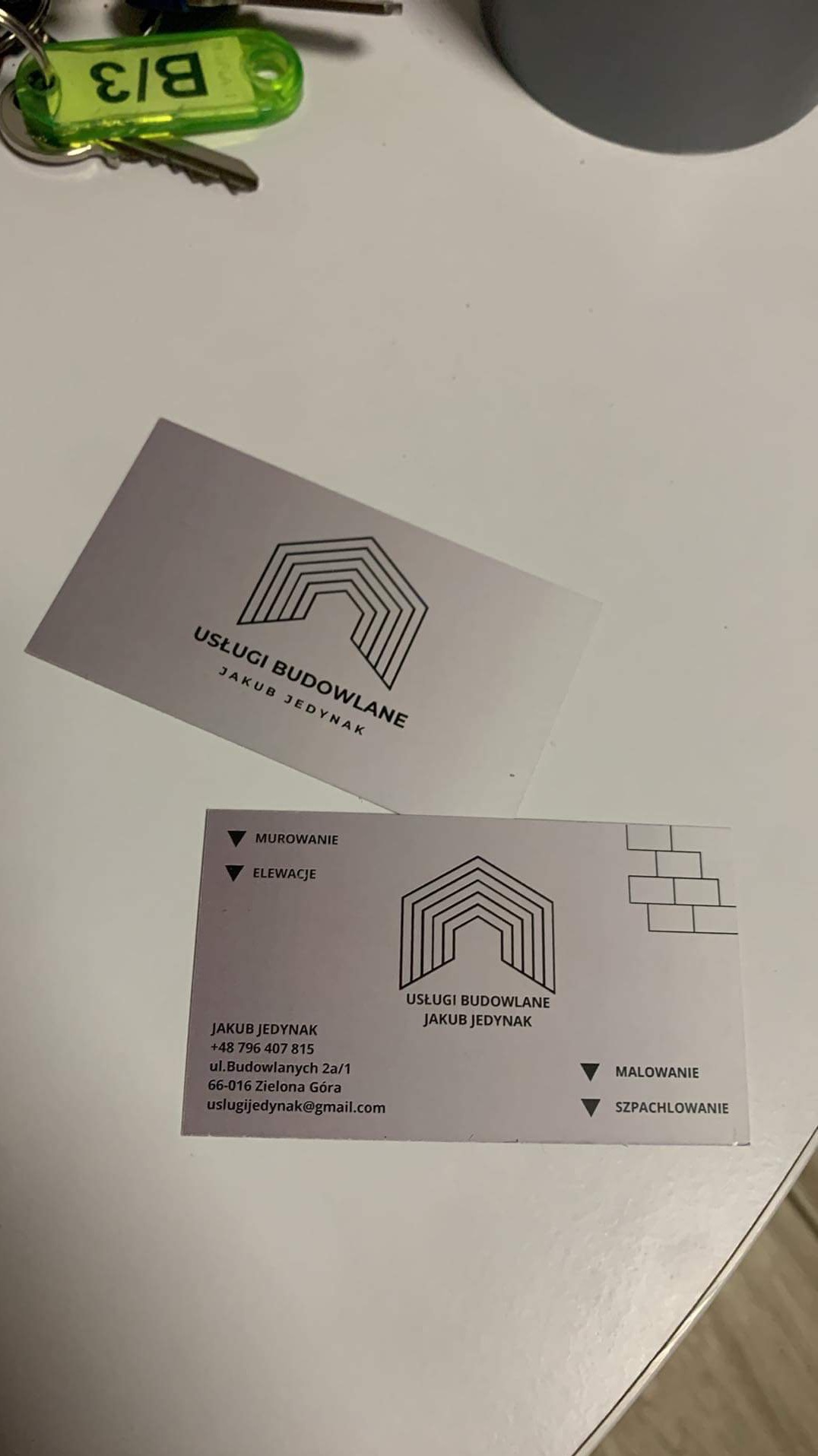 Dwie wizytówki firmy budowlanej Jakub Jedynak z adresem, numerem telefonu i zakresem usług: murowanie, elewacje, malowanie, szpachlowanie, leżące na białym tle obok kluczy i kubka.