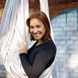 Uśmiechnięta kobieta w czarnym stroju, trzymająca się jasnego hamaka do aerial jogi na tle ceglanej ściany z graffiti.