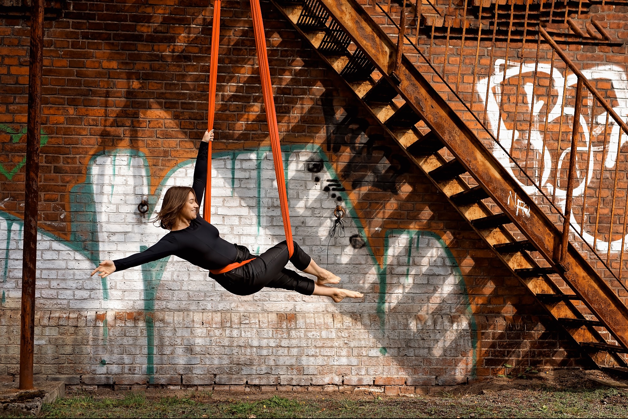Kobieta w czarnym stroju wykonuje ćwiczenie w powietrzu na pomarańczowych szarfach, na tle ceglanej ściany z graffiti i metalowych schodów przeciwpożarowych.