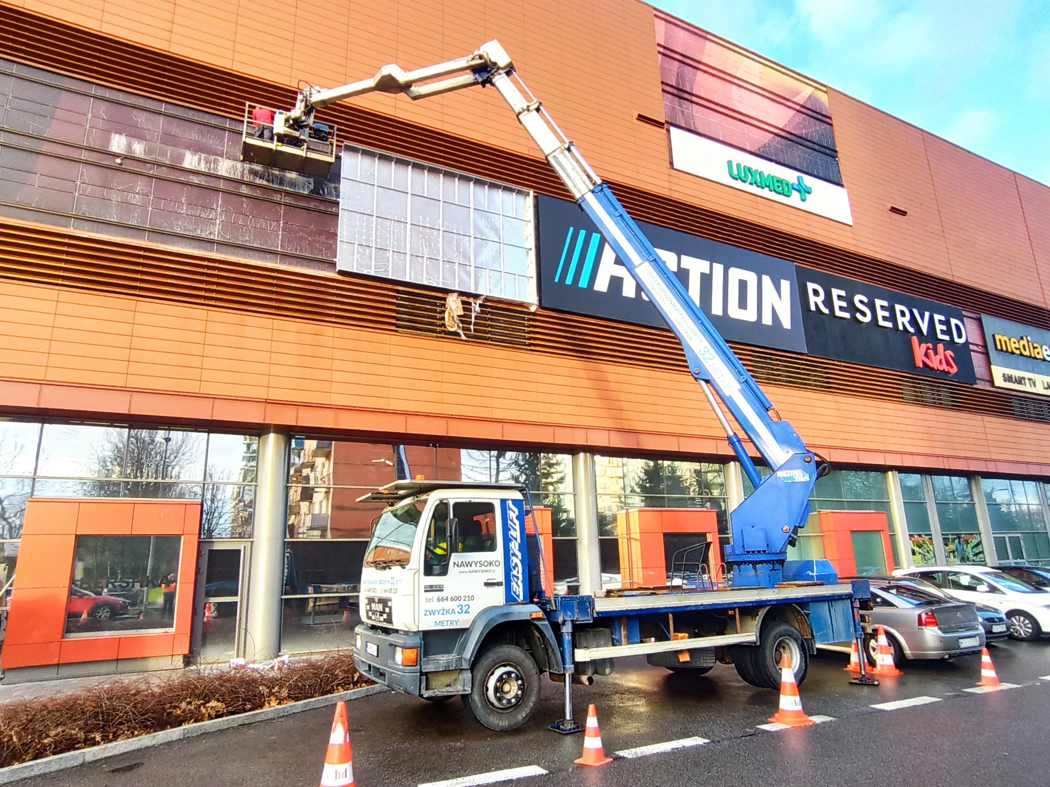 Czyszczenie elewacji budynku handlowego z wykorzystaniem platformy na samochodzie ciężarowym. Widoczne znaki firmowe na elewacji i pracownik w koszu platformy.