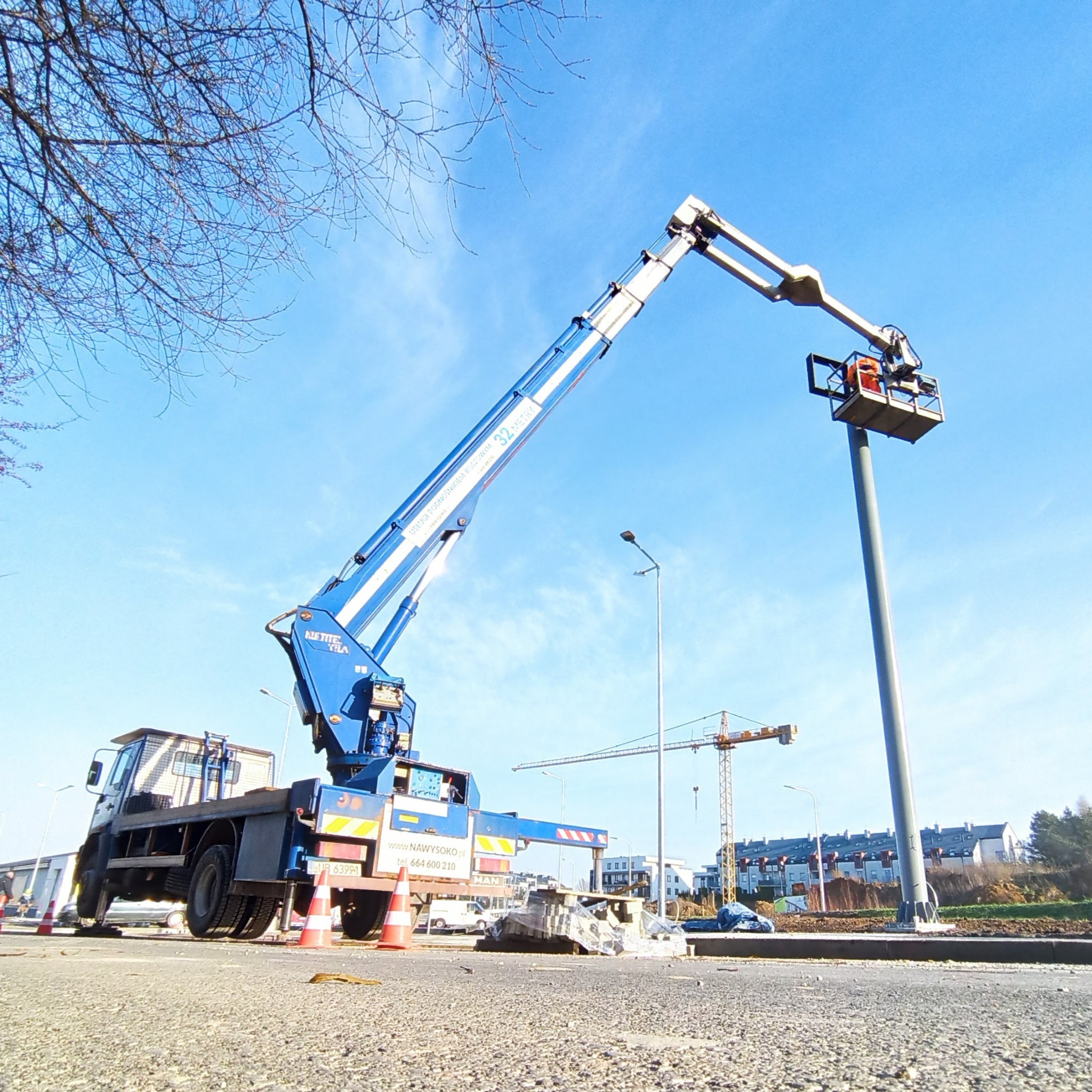 Pracownik w pomarańczowym kombinezonie w koszu podnośnika teleskopowego przy słupie oświetleniowym; w tle budowa i błękitne niebo.