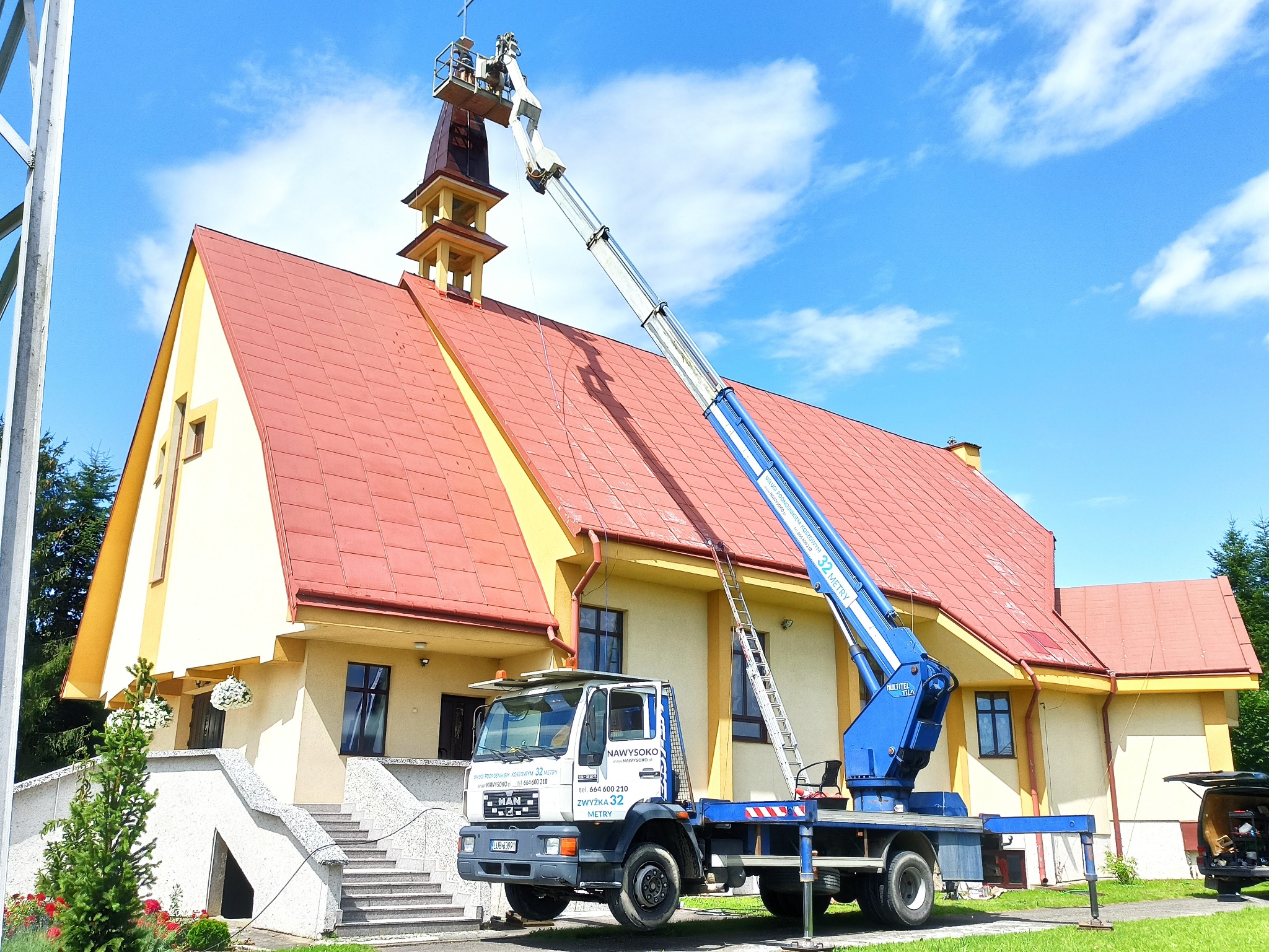 Niebieski podnośnik koszowy MAN z wysięgnikiem sięgającym wieży kościoła z czerwoną dachówką na tle błękitnego nieba.