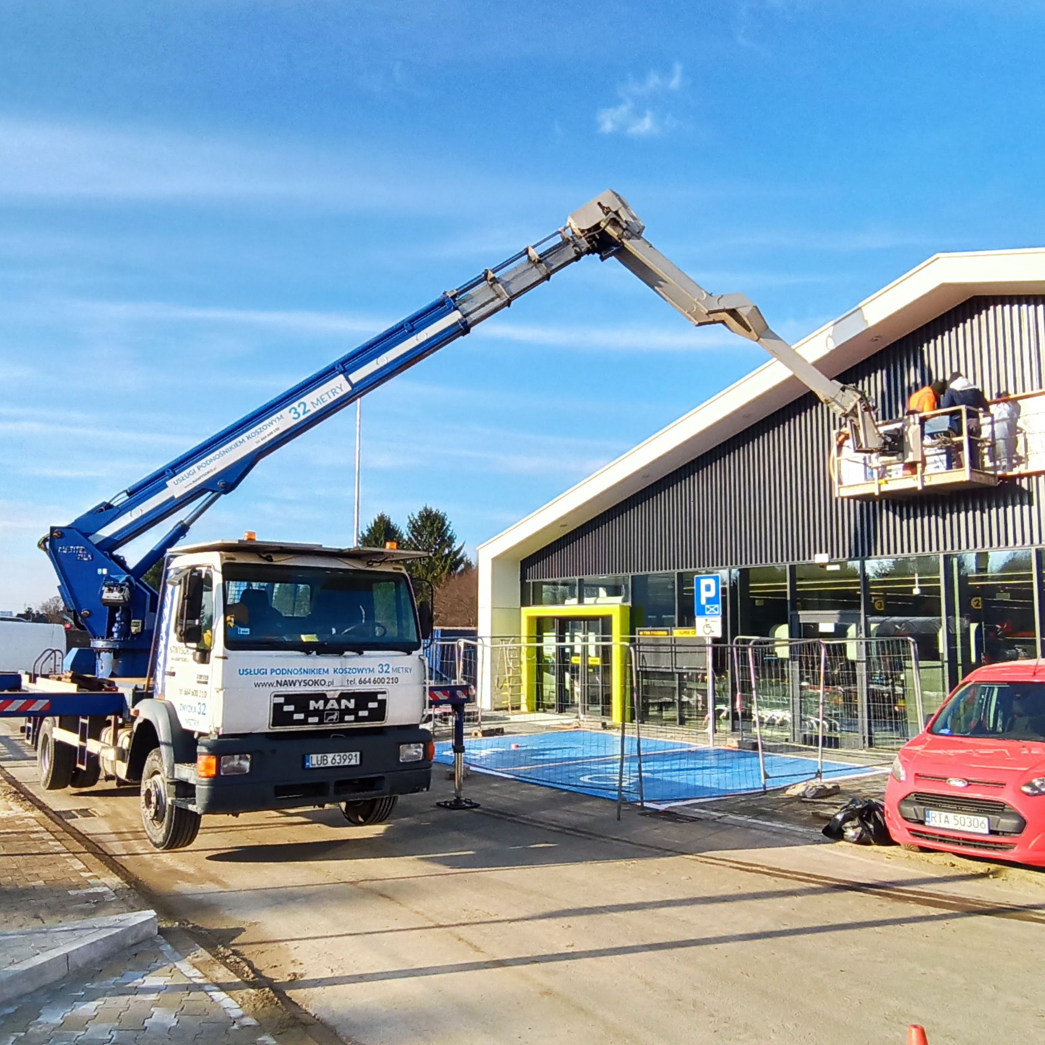 Biała ciężarówka z niebieskim wysięgnikiem platformy roboczej na tle budynku z czarnymi panelami, ekipa w koszu platformy wykonuje prace na wysokości.