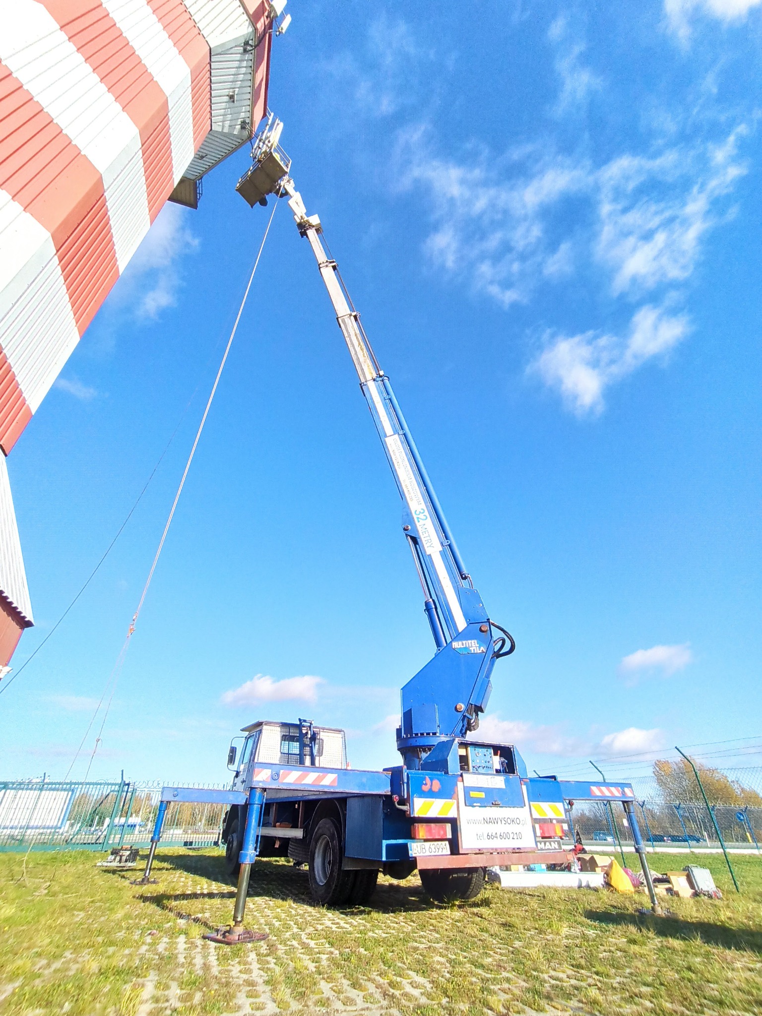 Niebieski pojazd z wysięgnikiem hydraulicznym sięga do górnej części wieży pomalowanej w czerwono-białe pasy na tle błękitnego nieba z chmurami.