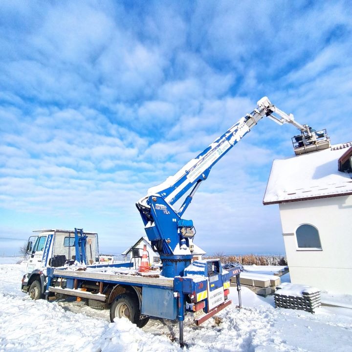 Niebieski podnośnik koszowy teleskopowy z białymi akcentami, stojący w śniegu, sięga do dachu białego, ośnieżonego domu z widocznym oknem łukowym.