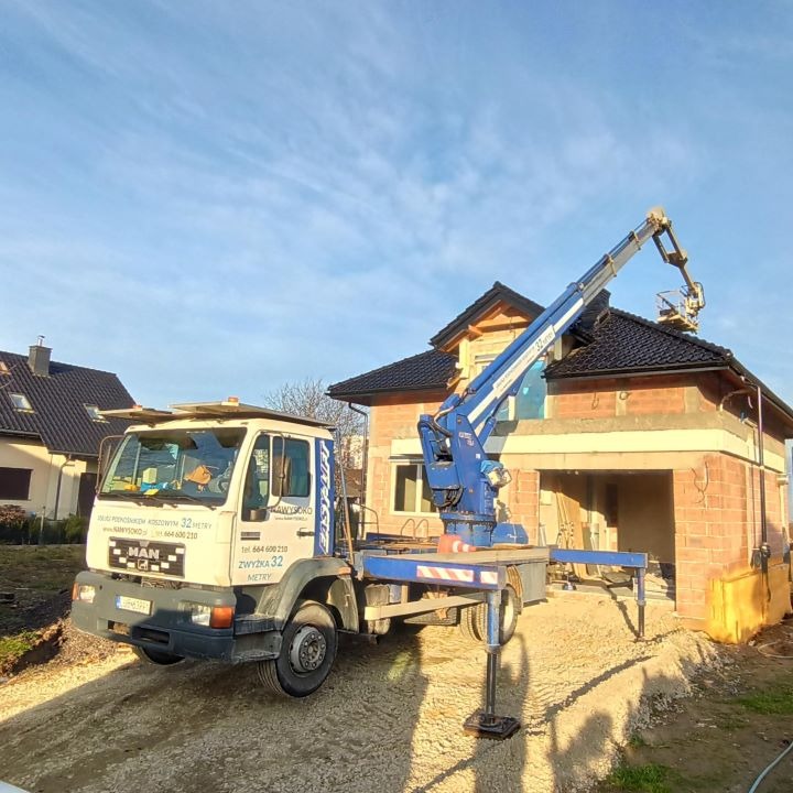 Biała ciężarówka z niebieskim ramieniem teleskopowym, platformą i napisem EASY LIFT, ustawiona przed budynkiem w trakcie budowy z czerwonej cegły i czarnym dachem. Platforma z pracownikiem...