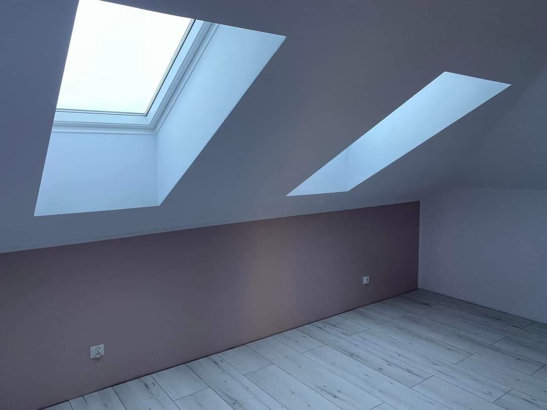 Poddasze po remoncie z dwoma oknami dachowymi, ściana pomalowana na dwa kolory: beżowy i biały, podłoga wyłożona panelami imitującymi drewno.