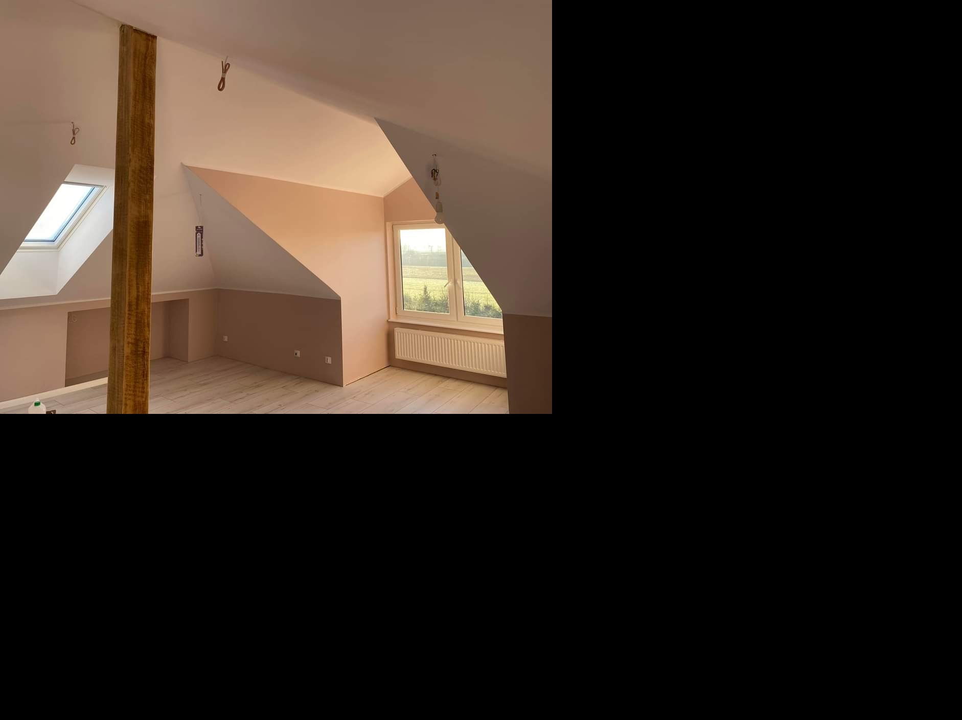 Poddasze po malowaniu, ściany pomalowane na dwa kolory, dolna część w odcieniach beżu, górna biała, widoczna drewniana belka, okno dachowe i grzejnik.