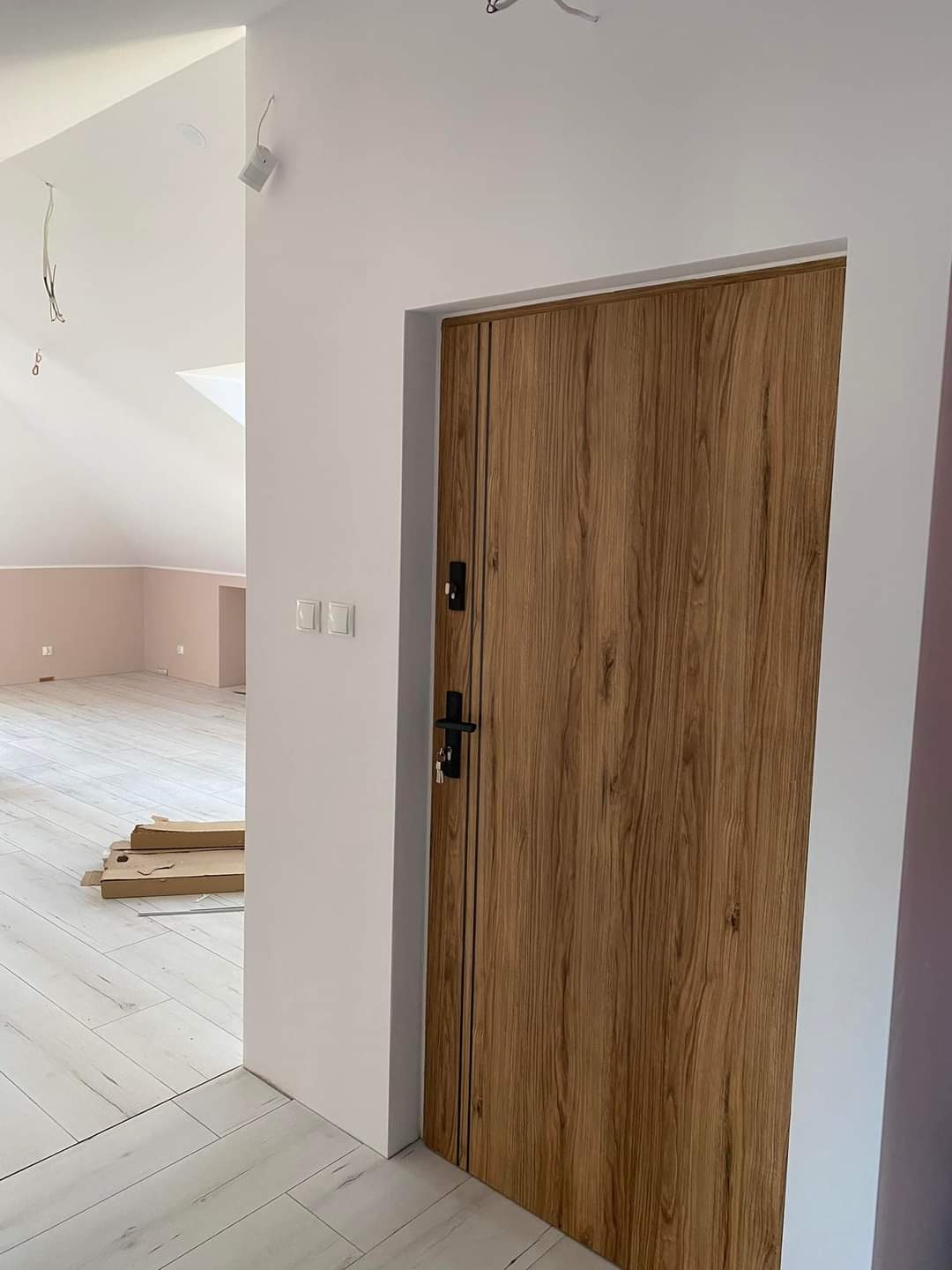 Zamknięte, nowoczesne drzwi wejściowe w odcieniach brązu z czarną klamką i dwoma pionowymi paskami, zamontowane w białej ścianie w nowym mieszkaniu z białymi panelami podłogowymi i widocznym...