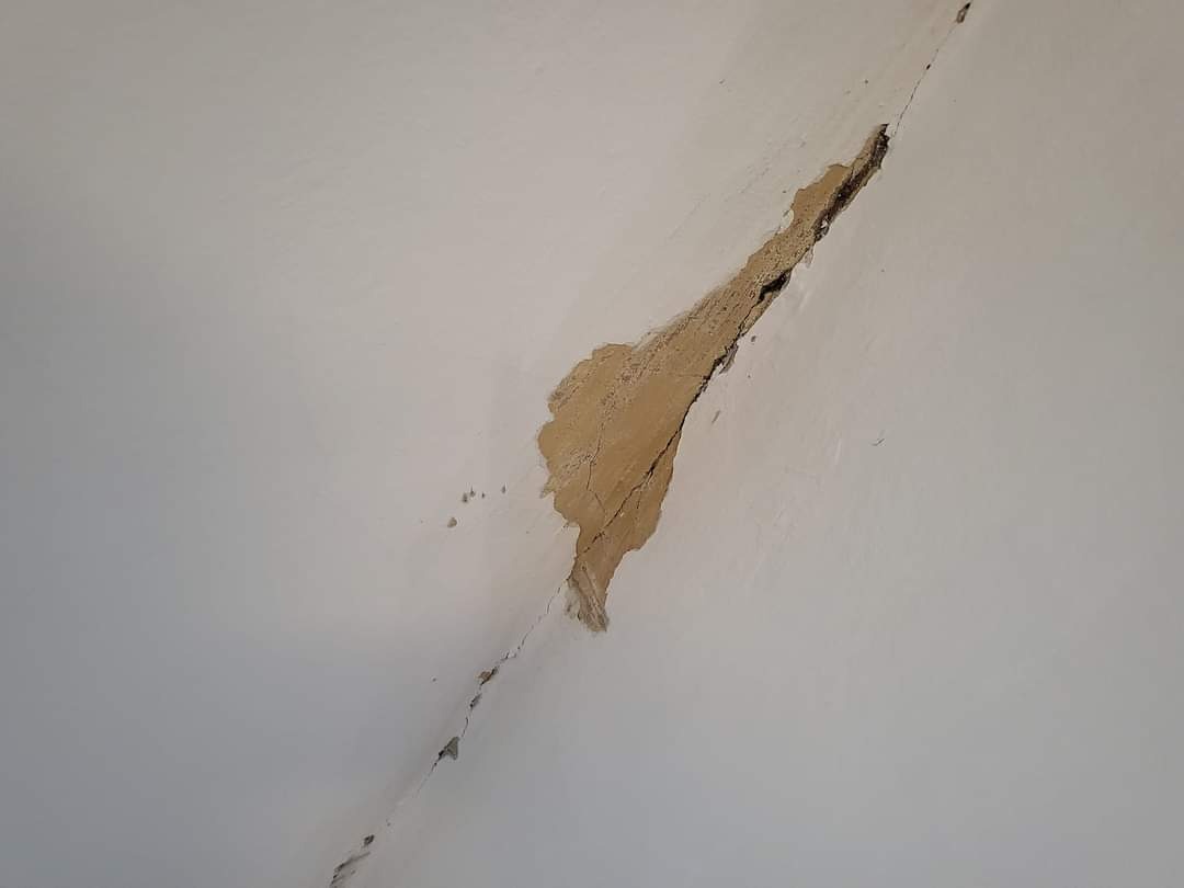 Pęknięcie i ubytek w pomalowanej na biało ścianie, odsłaniające warstwę podkładową i widoczne rysy, wskazujące na uszkodzenie struktury.