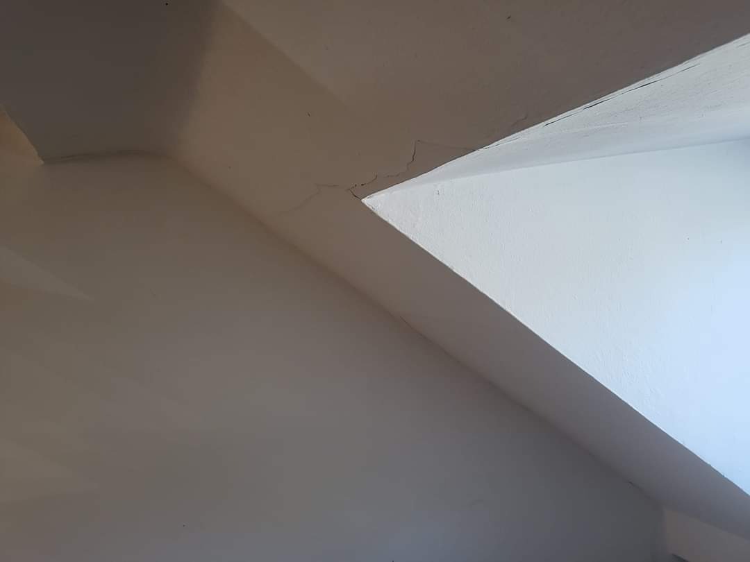 Białe ściany i sufit pod skosem z widocznymi pęknięciami i nierównościami, wymagające naprawy.