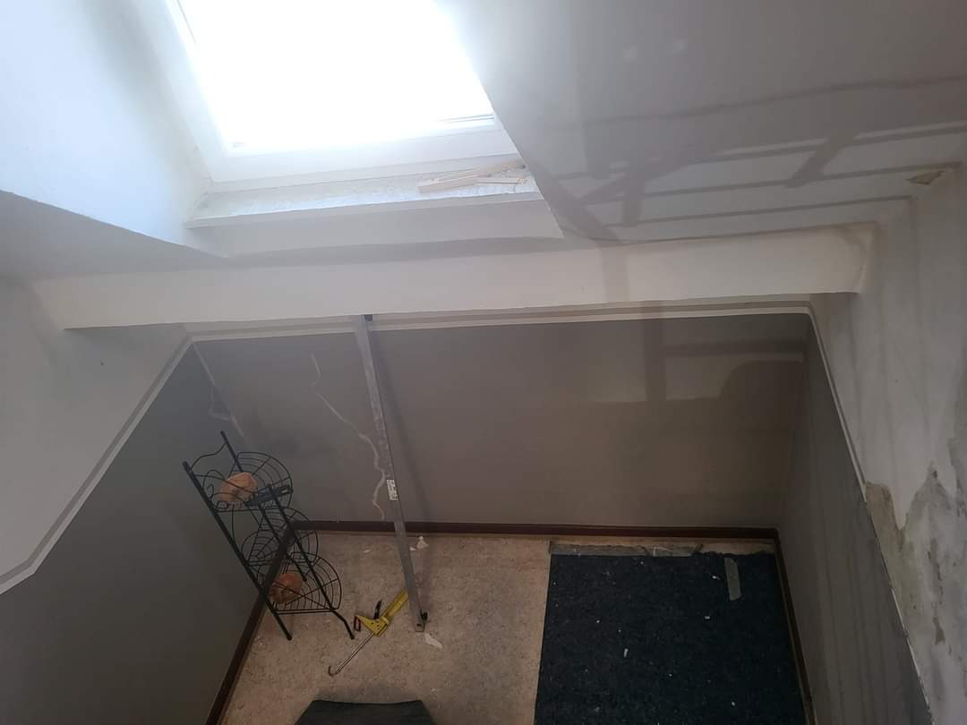 Wnętrze w trakcie remontu: ściany po szpachlowaniu, widoczny otwór okienny w suficie, metalowy regał na podłodze i poziomica oparta o ścianę.