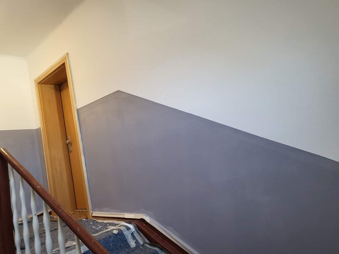 Klatka schodowa po malowaniu z dwukolorową ścianą w odcieniach szarości i bieli, zabezpieczone schody, widoczna drewniana ościeżnica drzwi.