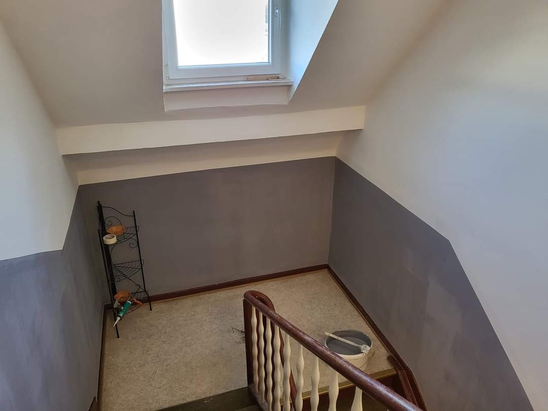 Klatka schodowa po częściowym malowaniu na szaro, widoczny fragment balustrady, wiadro z farbą i drabinka z narzędziami przy ścianie.