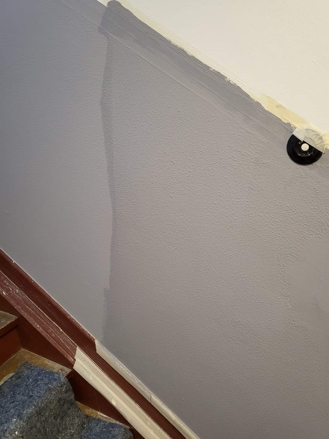 Świeżo pomalowana szara ściana przy schodach, widoczna taśma malarska zabezpieczająca listwy przypodłogowe i oprawę oświetleniową na suficie.