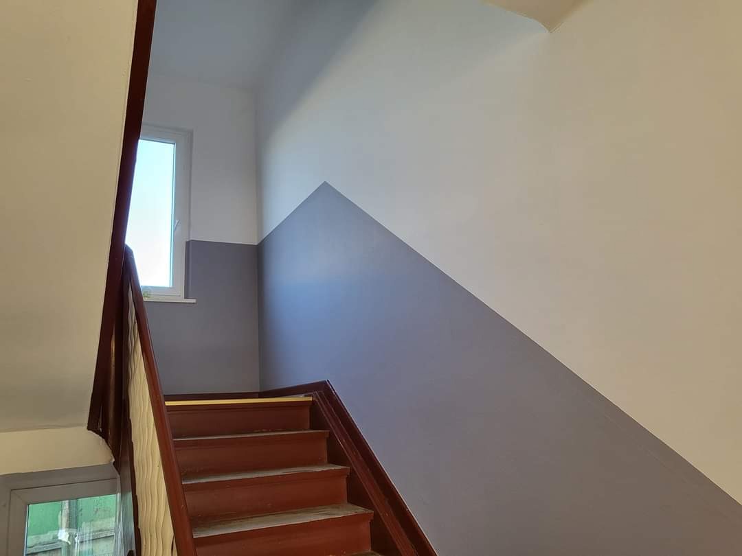 Klatka schodowa po malowaniu: ściany pomalowane na biało i szaro, widoczne drewniane schody i okno.