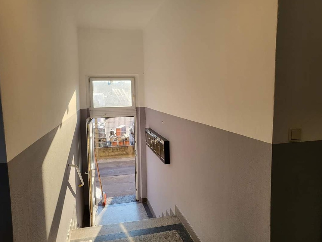 Klatka schodowa po odświeżeniu, z dwukolorowymi ścianami (górna część biała, dolna szara), widoczne skrzynki pocztowe i otwarte drzwi wejściowe z refleksami światła słonecznego.