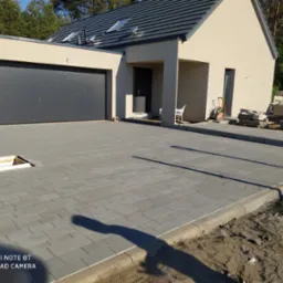 Świeżo ułożona szara kostka brukowa na podjeździe do domu w Gryficach, z widocznym otworem rewizyjnym i przygotowanym obrzeżem, na tle nowoczesnego domu z garażem.