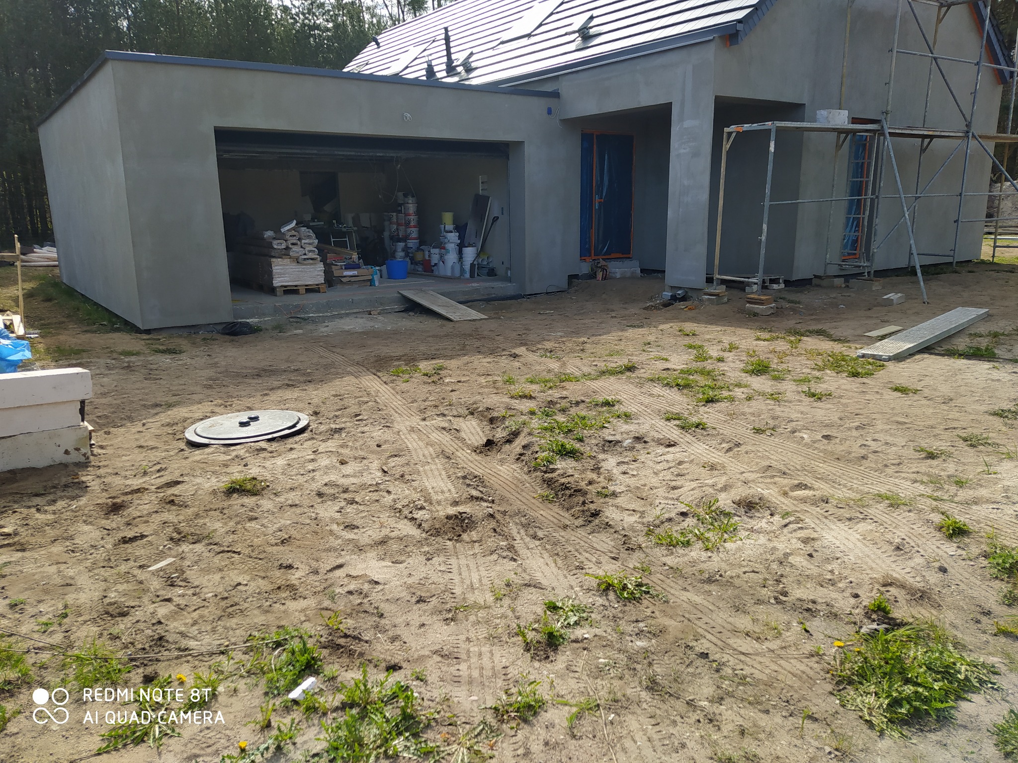 Nowo otynkowany dom jednorodzinny z garażem w trakcie wykończenia, widoczne rusztowania i materiały budowlane, teren wokół wyrównany z widocznymi śladami opon.