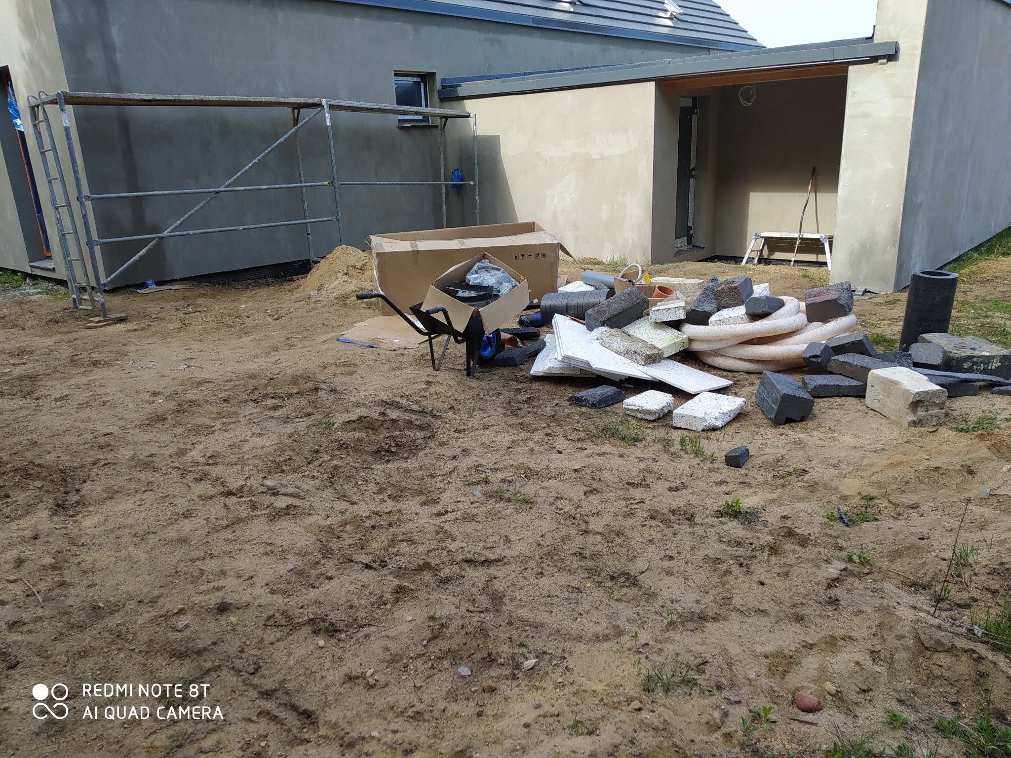 Teren budowy z rozrzuconymi materiałami: kostki brukowe, rury drenarskie, styropian, kartony i taczka, na tle surowego budynku z rusztowaniem.