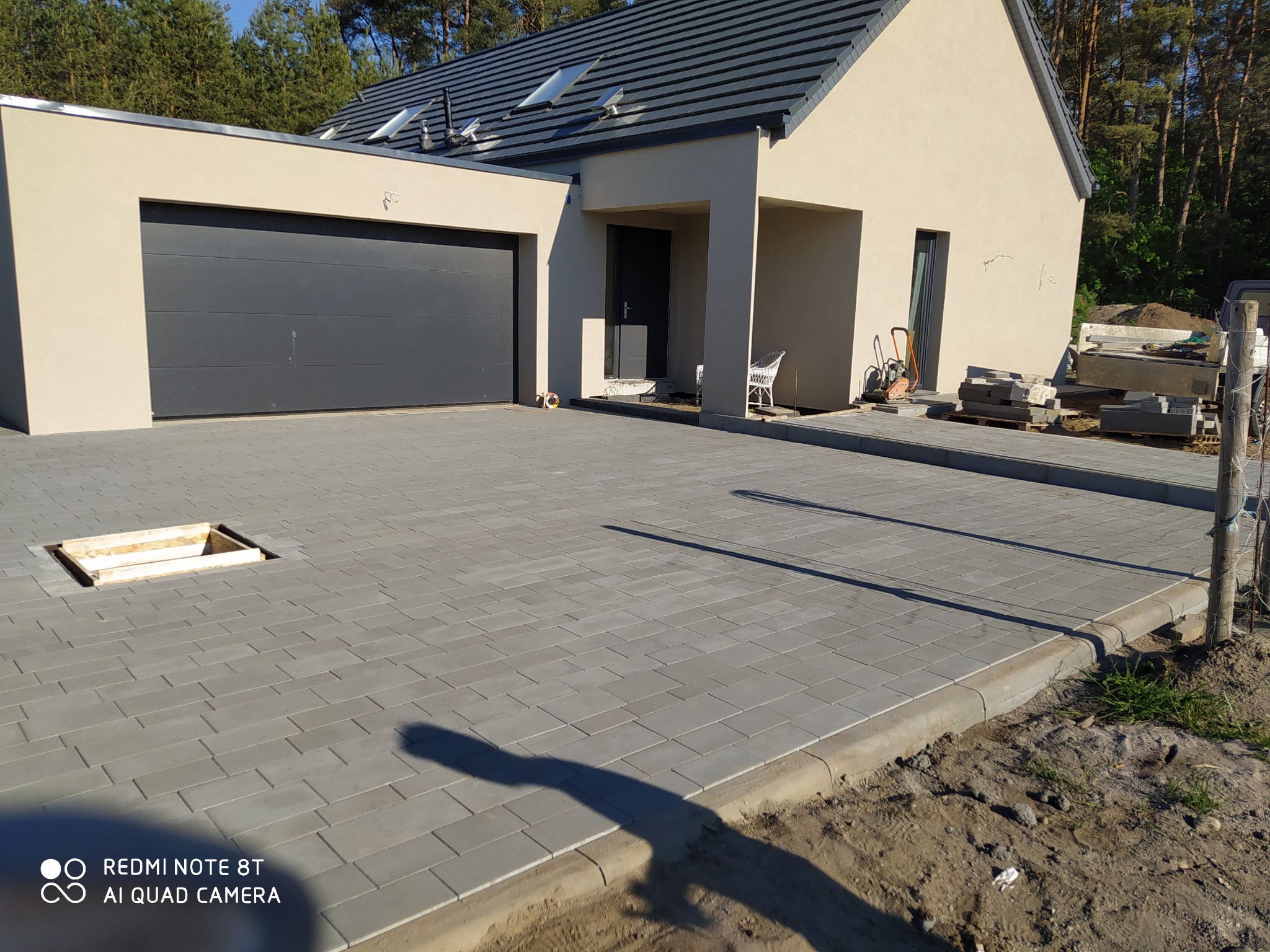 Świeżo ułożona szara kostka brukowa na podjeździe do domu w Gryficach, z widocznym otworem rewizyjnym i przygotowanym obrzeżem, na tle nowoczesnego domu z garażem.