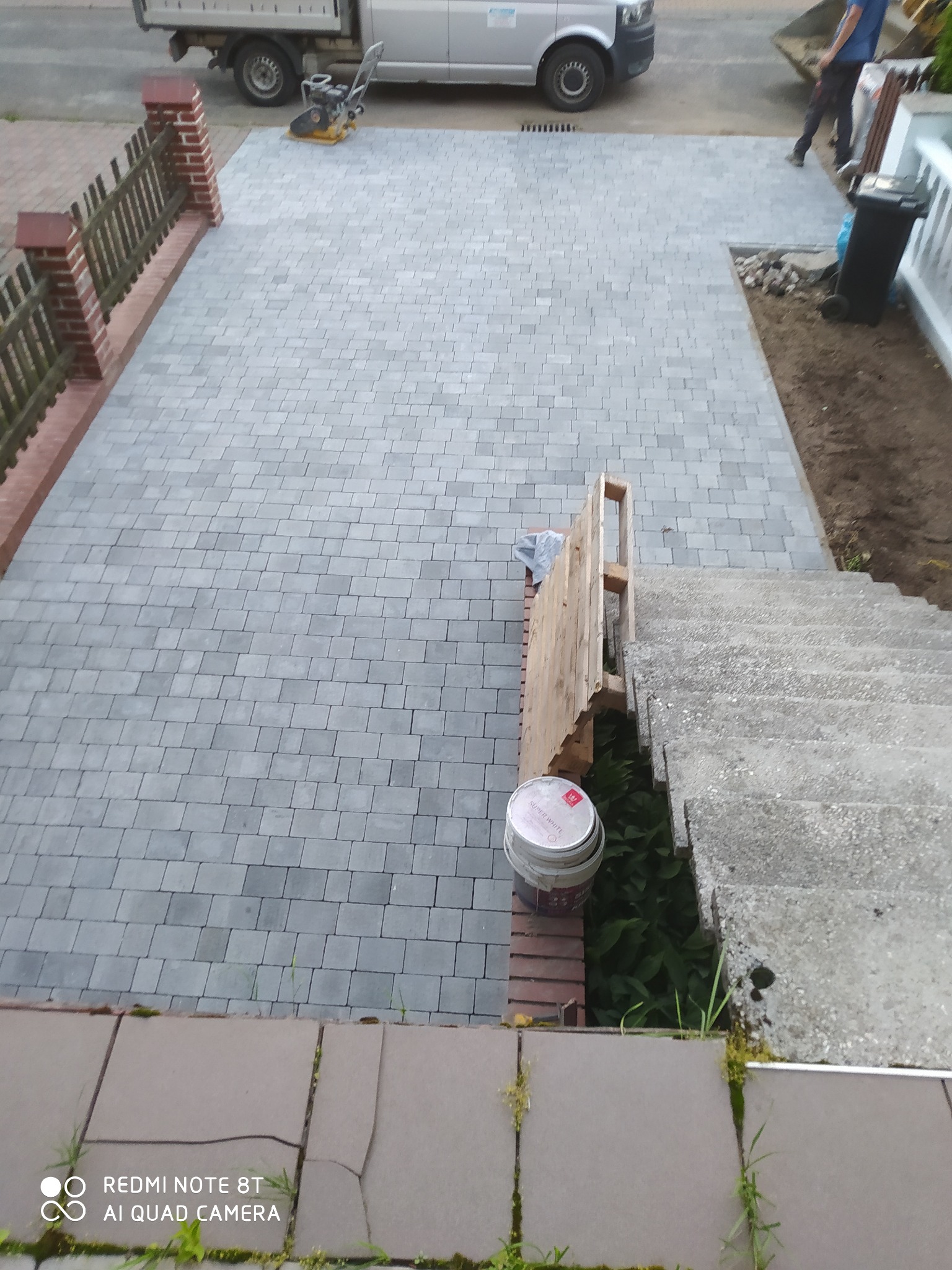 Świeżo ułożona szara kostka brukowa na podjeździe, widoczna ubijarka wibracyjna, częściowo ogrodzenie z ceglanymi słupkami, paleta oparta o schody i wiaderko farby.
