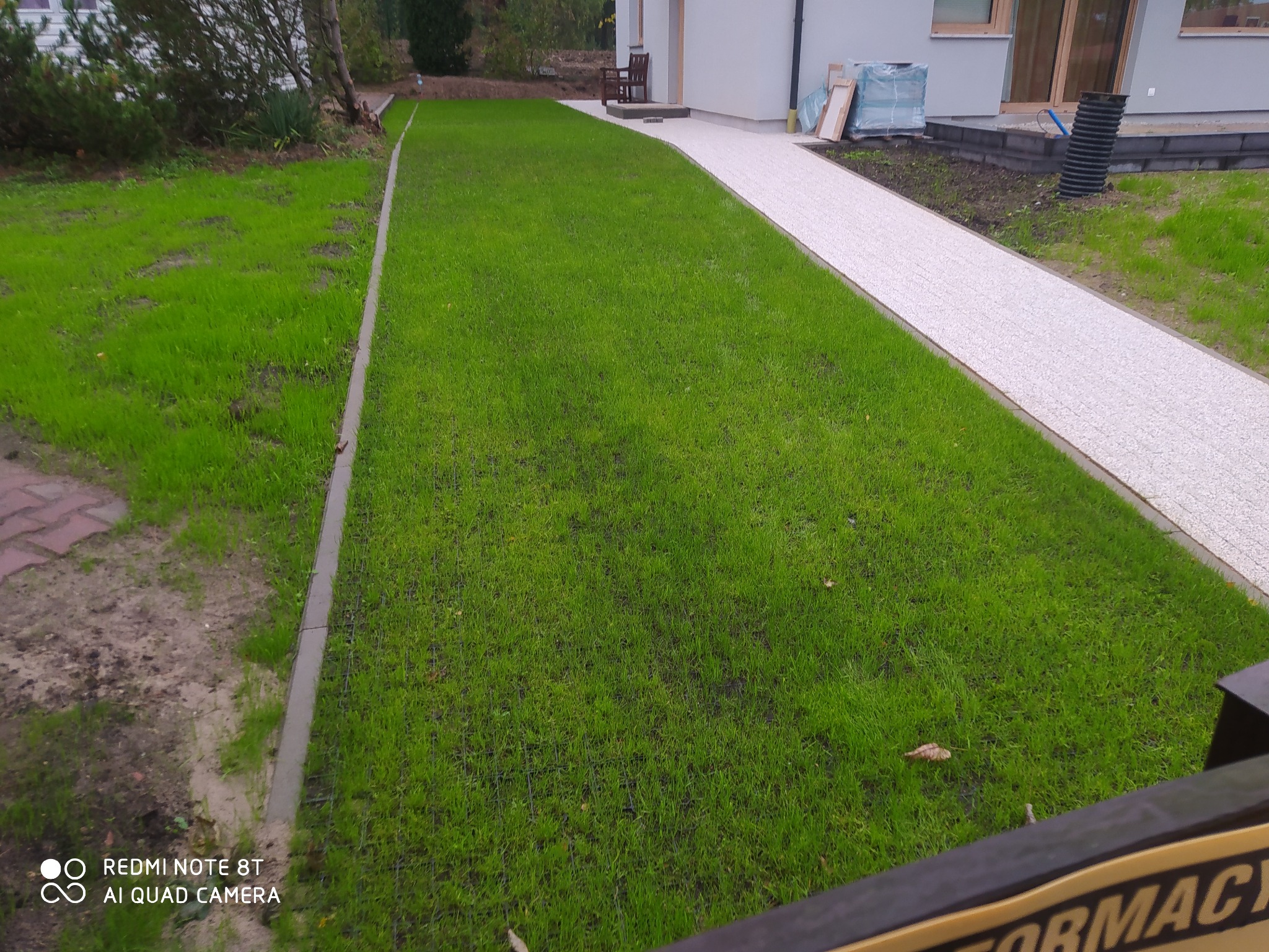 Świeżo założony trawnik z siatką wzmacniającą, obrzeża betonowe, ścieżka z białego kruszywa prowadząca do domu, widoczne elementy drenażu przy budynku.